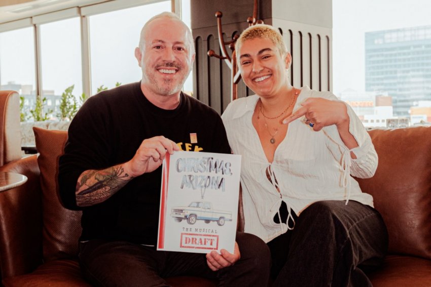 Dan Clapson and co-creator Zenon smiling on a leather sofa while holding a draft copy of the script for Christmas, Arizona The Musical.