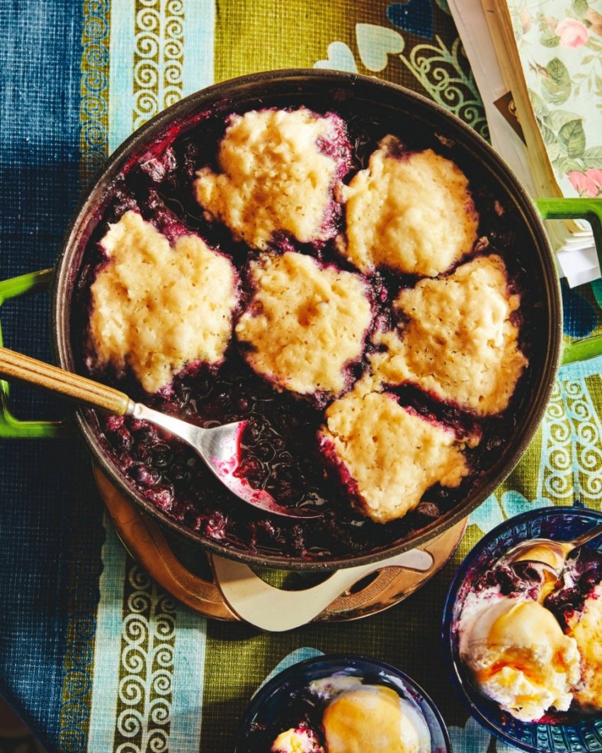 A portrait photo of a completed dish of jammy blueberries and lemon dumplings served with vanilla ice cream and a drizzle of maple syrup.